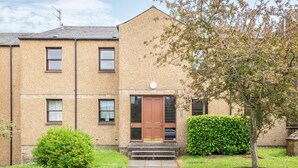A two-story building with a brown door and windows, a small garden with a tree, and a clear sky. - Kate Kennedy Court (No 2) (St Andrews)