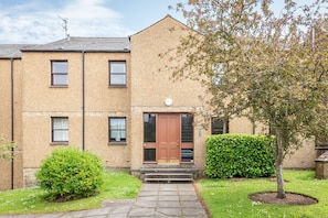 A two-story building with a brown door and windows, a small garden with a tree, and a clear sky. - Kate Kennedy Court (No 2) (St Andrews)