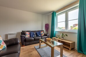 Ein modernes Wohnzimmer mit einer Ledersofa, einem Glastisch und Blick ins Freie durch ein großes Fenster mit türkisfarbenen Vorhängen.