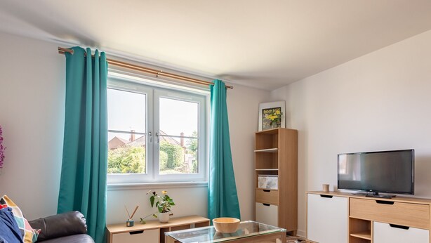 A living room with a sofa, a coffee table, a TV on a stand, a bookshelf, and a window with curtains. - Kate Kennedy Court (No 2) (St Andrews)