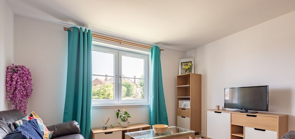 Ein Wohnzimmer mit einer Couch, einem Glastisch, einem Fernseher auf einem Holzgestell, einem Bücherregal und einem Fenster mit türkisfarbenen Vorhängen.