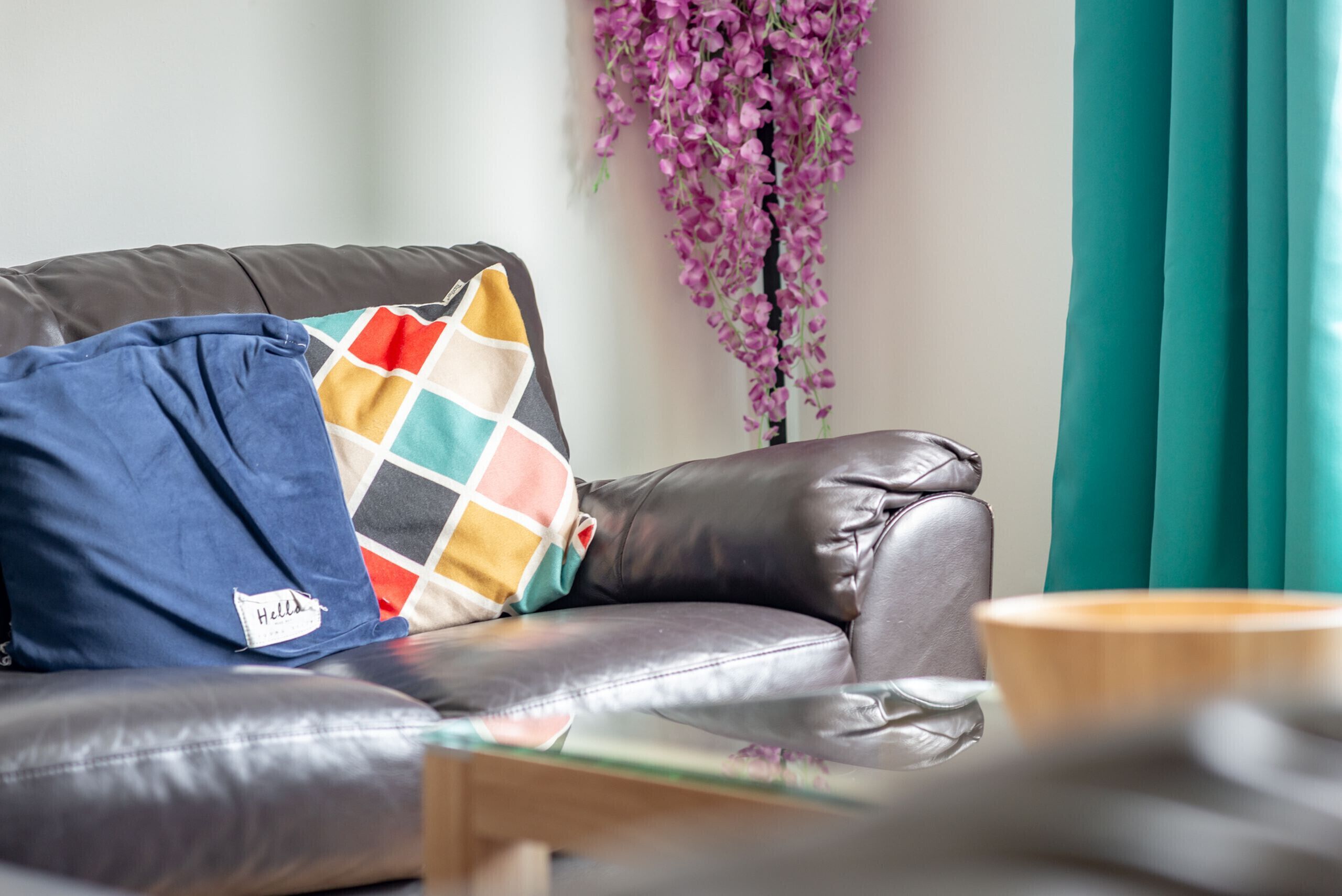 A leather sofa with a colorful throw pillow and a "Hello" tag, a wooden side table, and a hanging purple flower arrangement.