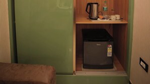 Una stanza compatta con una porta scorrevole verde, un piccolo ripiano in legno con un bollitore, una bottiglia d'acqua e due tazze, e un frigorifero compatto.