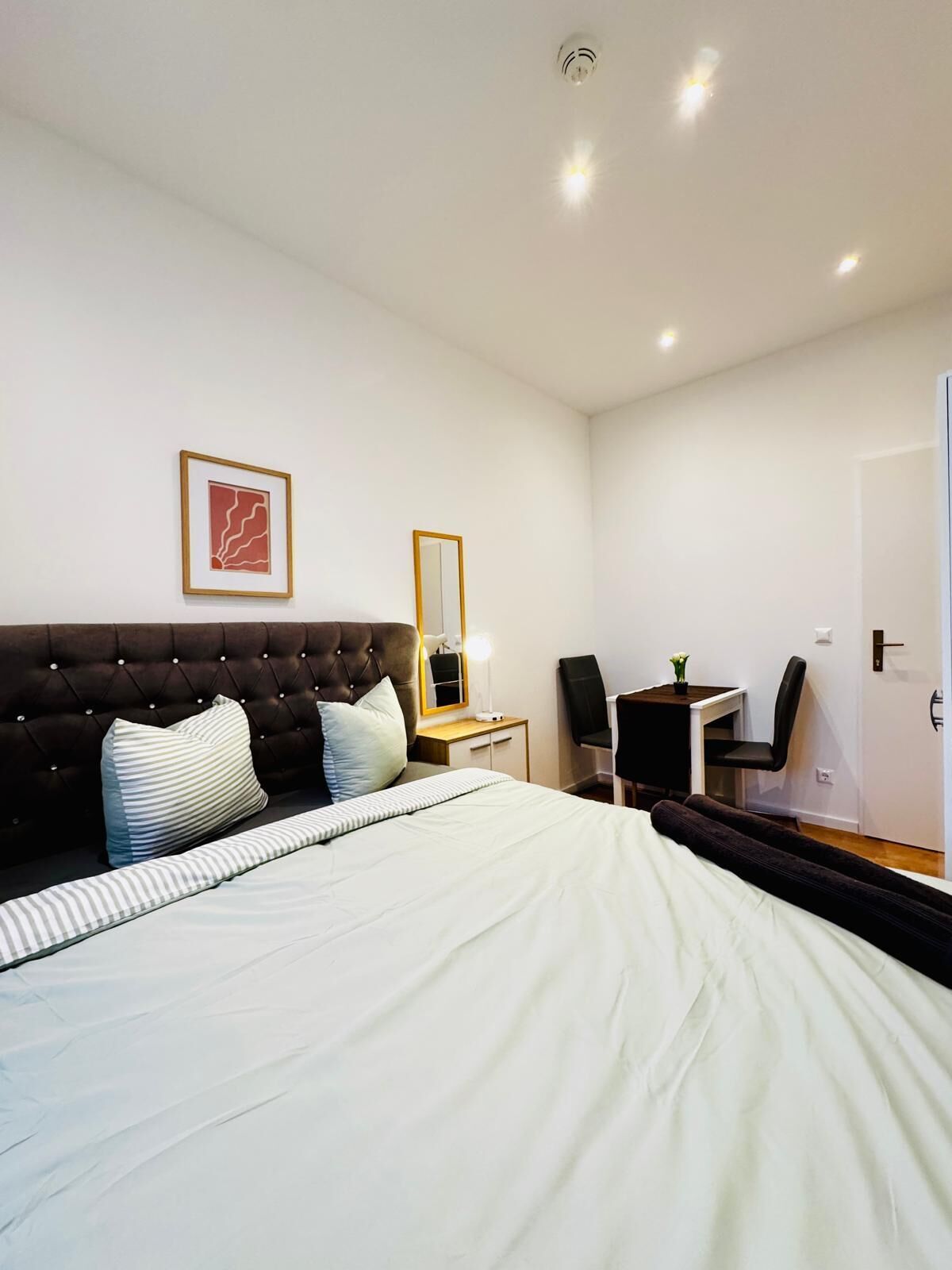 A bedroom with a large bed, a headboard, a bedside table with a lamp, a chair, a desk, and a mirror.