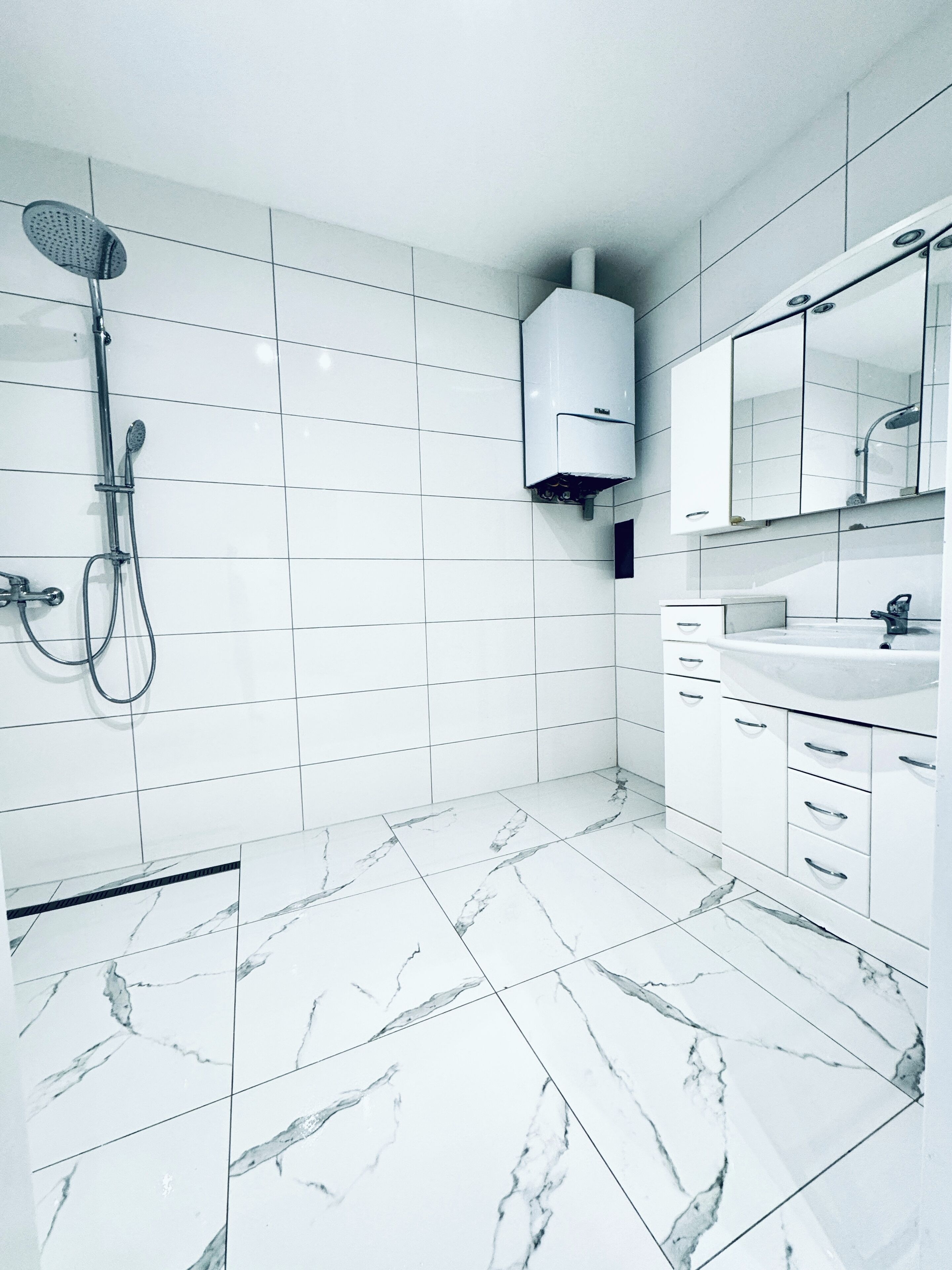 A modern bathroom with white tiles, a marble floor, a shower, a sink, and a mirror.