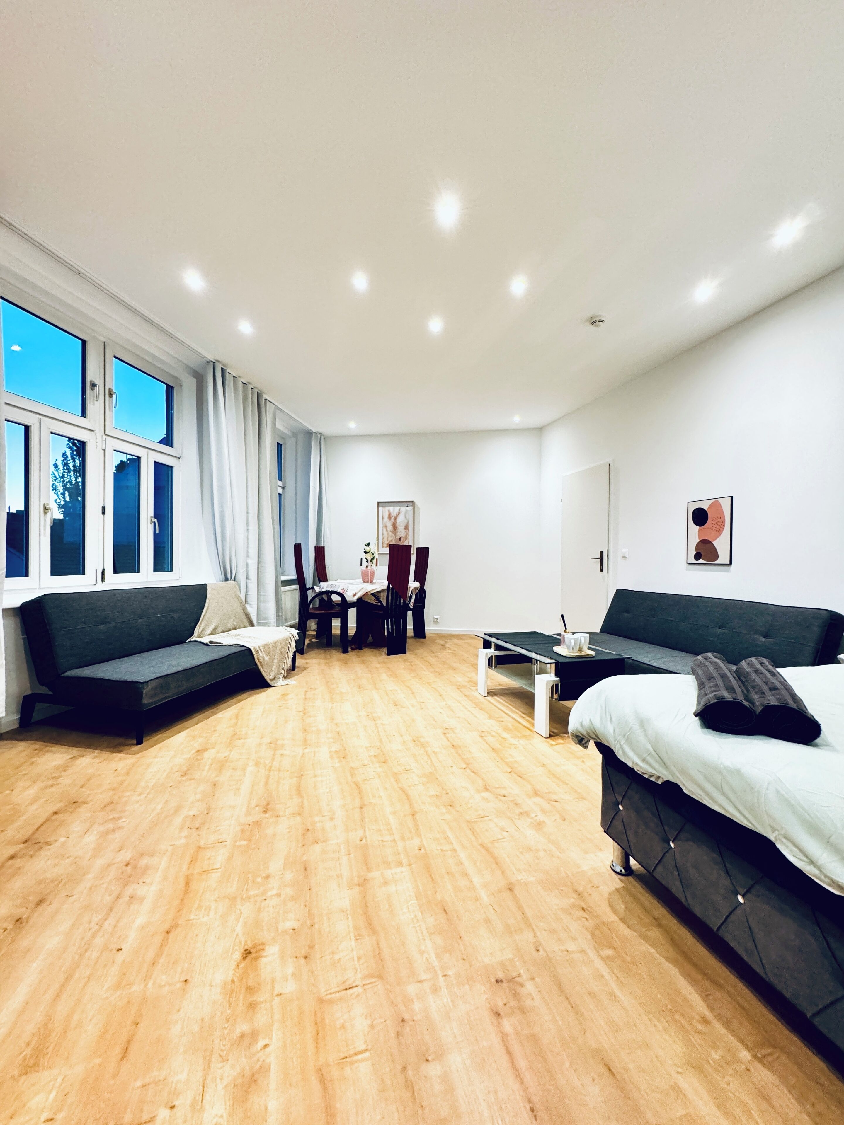 Una habitación moderna y luminosa con suelo de madera, un sofá, una cama, una mesa de comedor con sillas y ventanas amplias.