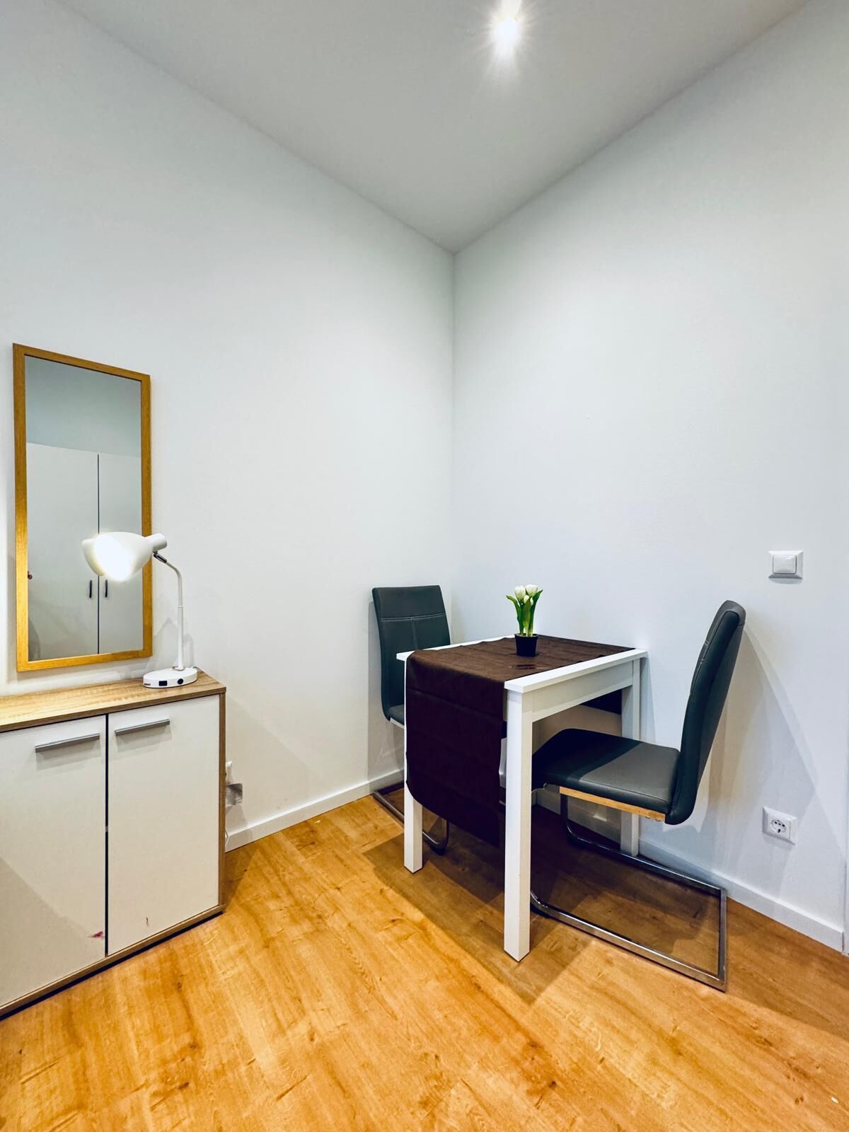 A small, clean room with a wooden floor, a white desk with a chair, a mirror, and a small table with a plant.
