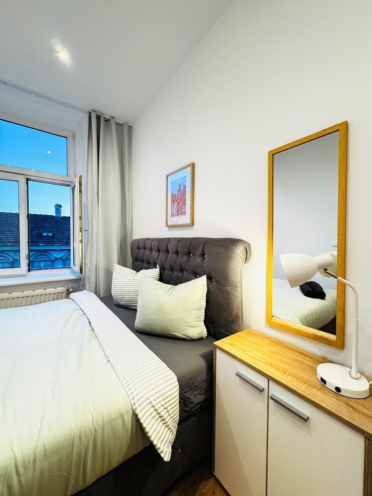 A modern bedroom with a large bed, a bedside table with a lamp, a mirror, and a view of the cityscape through a window.