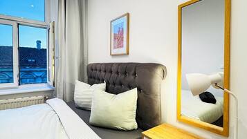 A modern bedroom with a large bed, a bedside table with a lamp, a mirror, and a view of the cityscape through a window.