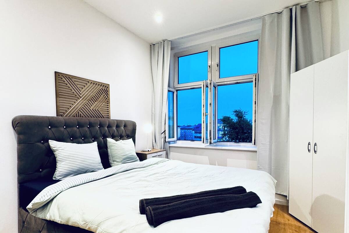 A modern bedroom with a large bed, a wardrobe, and a window with curtains.