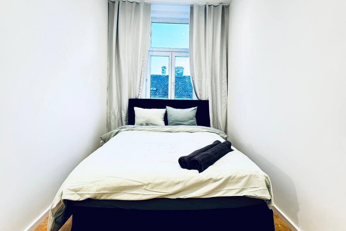 A small, modern bedroom with a single bed, white curtains, and a view of buildings outside.