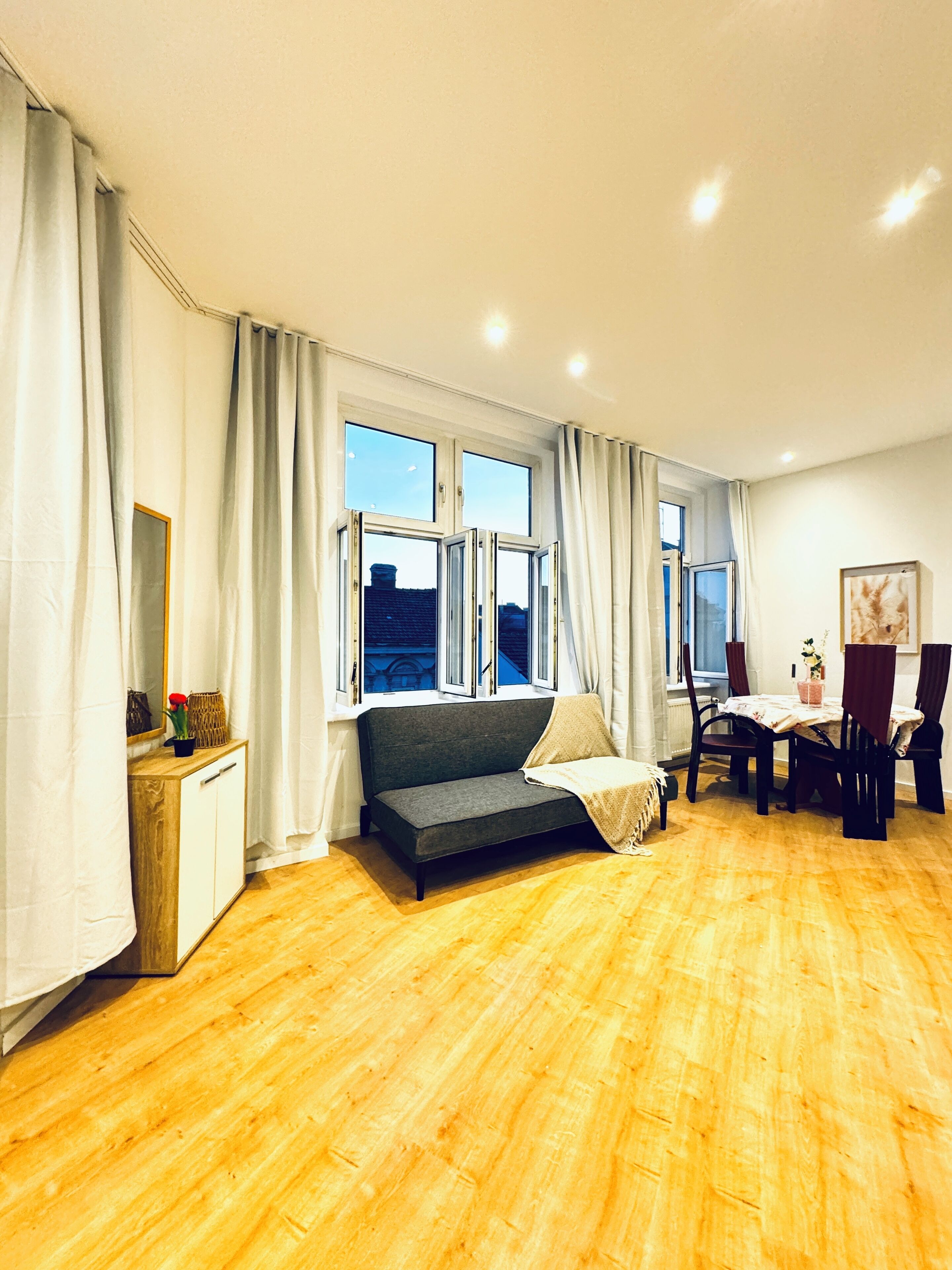 Een ruime kamer met een houten vloer, een grijze bank, een eettafel met stoelen en een raam met gordijnen.