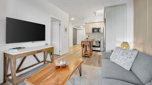 Een moderne woonkamer met een flatscreentelevisie, een houten salontafel, een grijze bank en een keuken op de achtergrond.