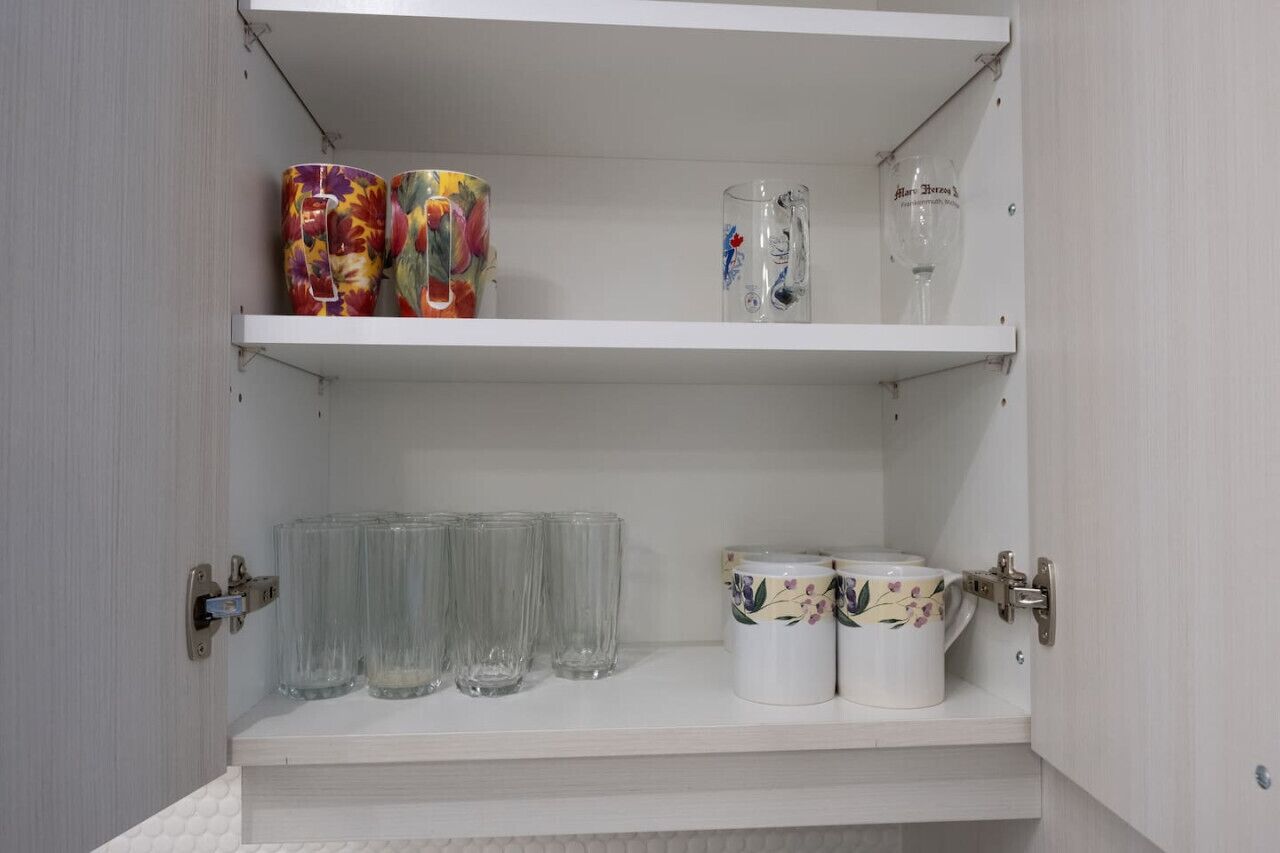 En vit hylla med dekorativa muggar, genomskinliga glas och ett vinglas.