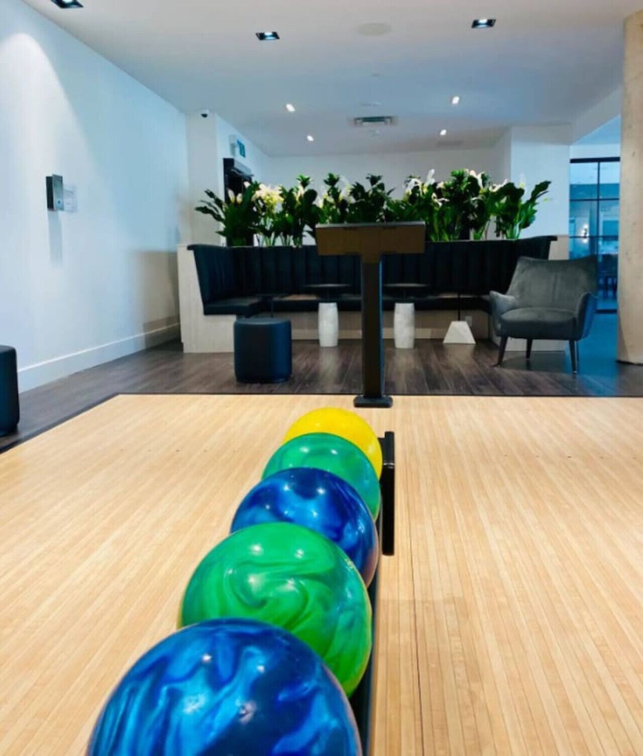 Una bolera con suelo de madera, una fila de coloridas bolas de bolos y una silla negra al fondo.