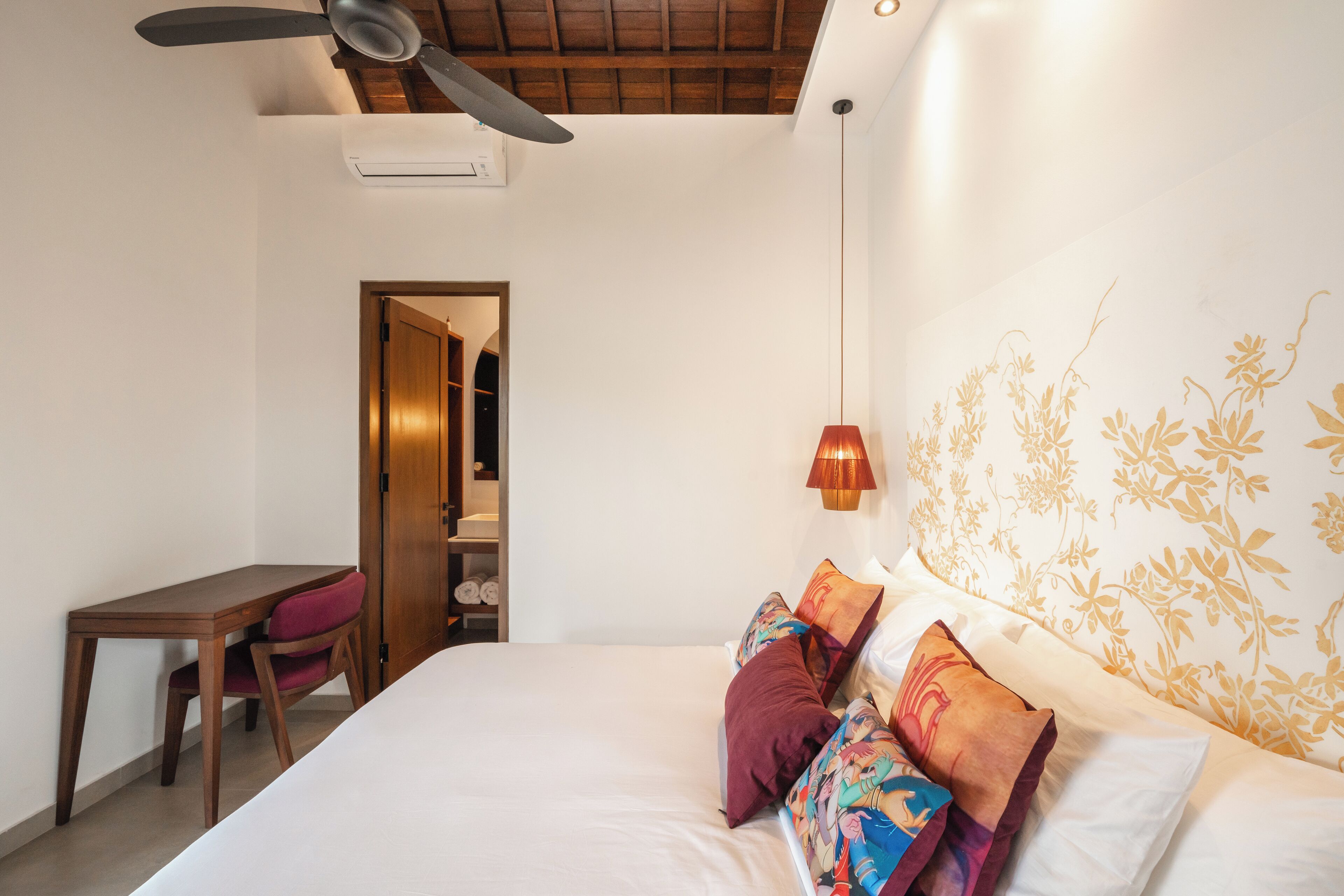 Ein Schlafzimmer mit einem Bett, einem Schreibtisch und einem Stuhl. Es gibt einen Deckenventilator, eine Klimaanlage und eine Wand mit floralem Muster.