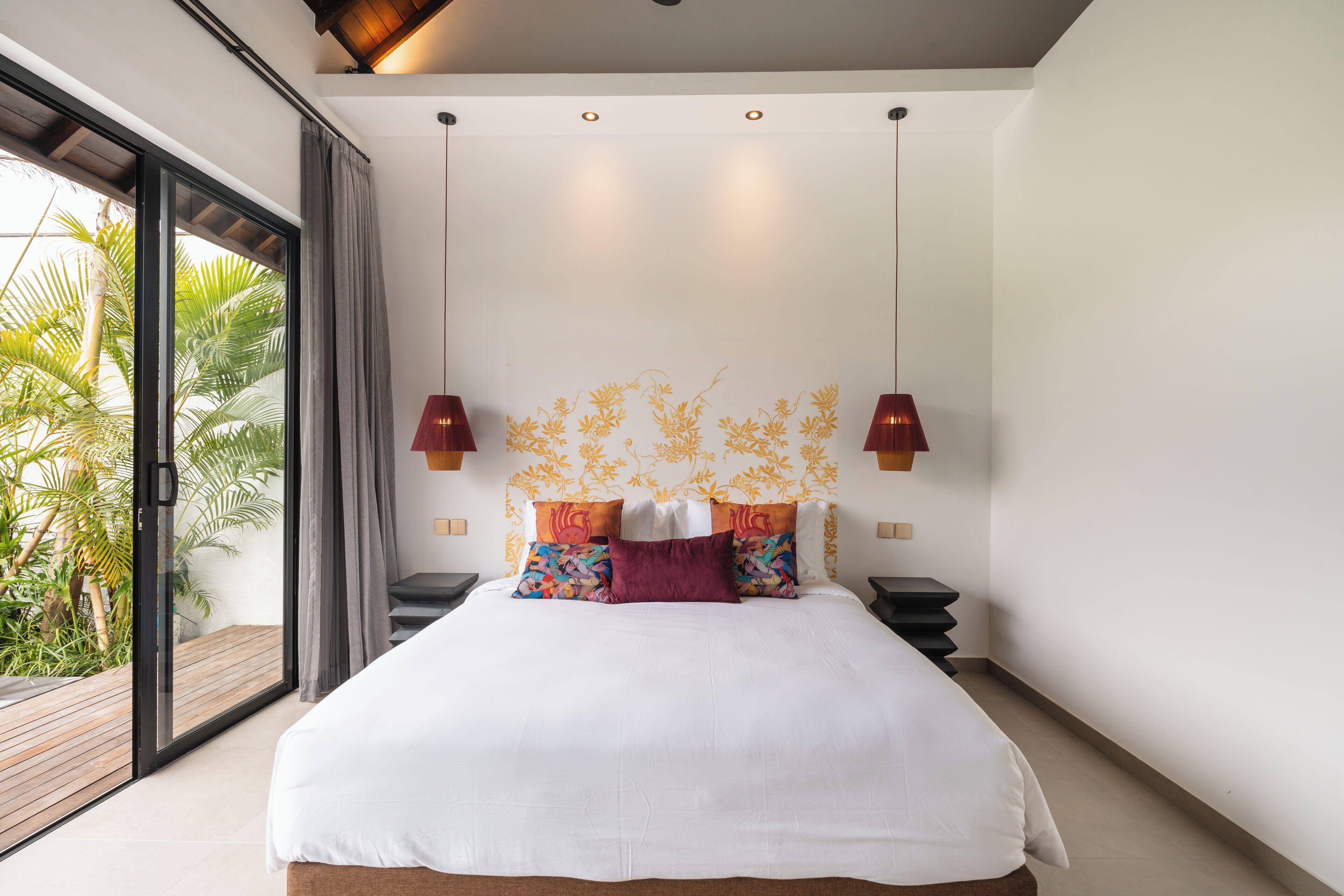 A modern bedroom with a large bed, a view of palm trees, and a ceiling with pendant lights.