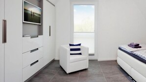 A modern bedroom with a white wardrobe, a white armchair with a striped pillow, a bed with a striped duvet, and a large window with a view of trees. - Ferienhaus am Yachthafen in Peenemuende (Peenemünde)