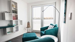 A modern living room with teal furniture, a white bookshelf, and a large window with a view. - Ferienhaus am Yachthafen in Peenemuende (Peenemünde)