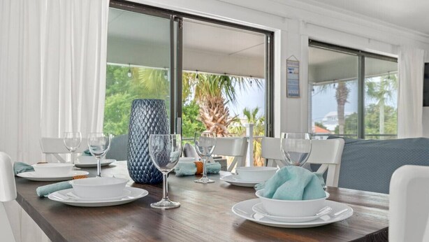 Un coin repas avec une table en bois dressée pour quatre personnes, des chaises blanches et une grande fenêtre donnant sur des palmiers.