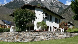 A two-story house with a stone wall in front, a mountain backdrop, and a clear sky.