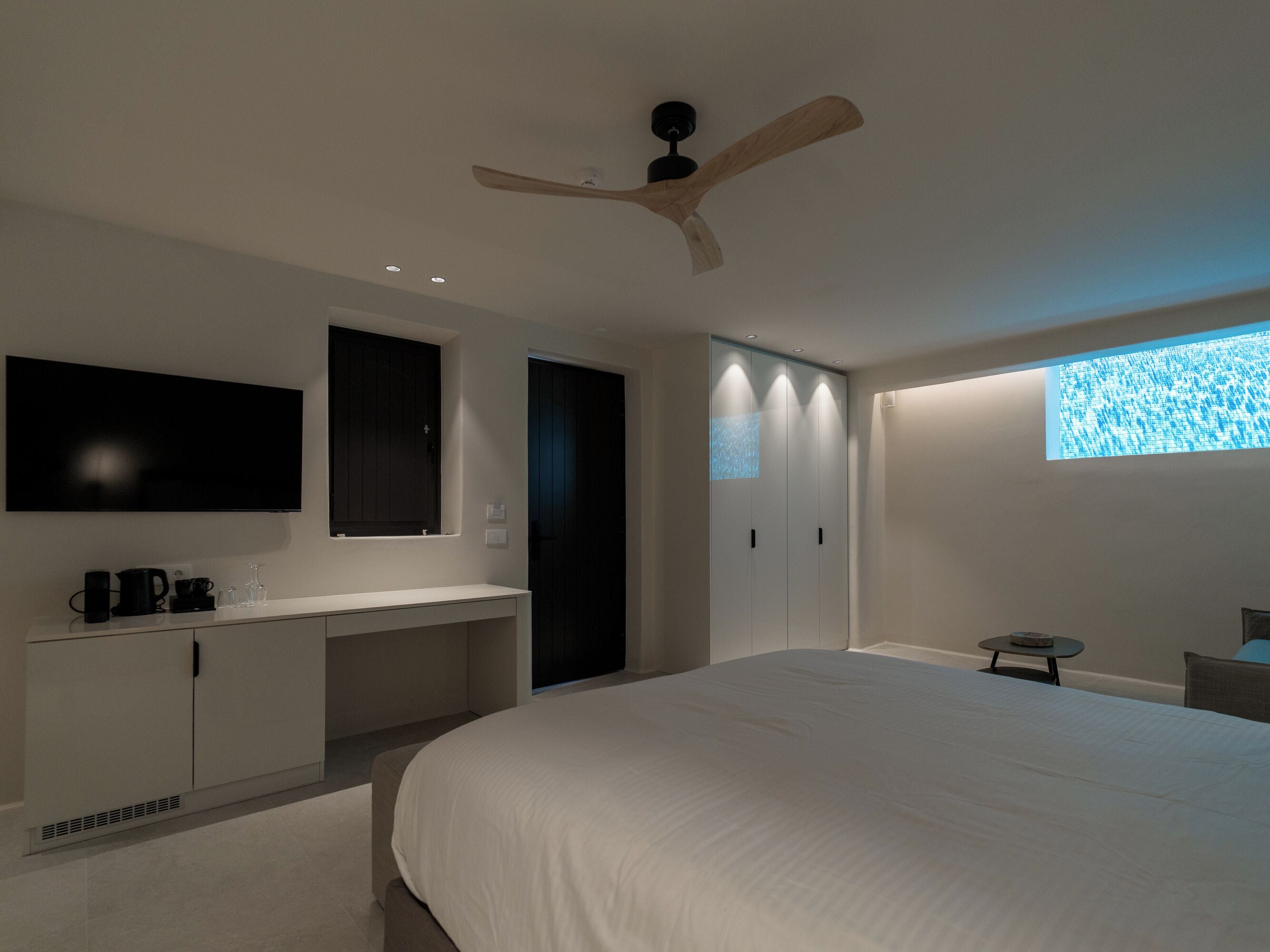 A modern bedroom with a large bed, a built-in desk, a ceiling fan, and a TV mounted on the wall.