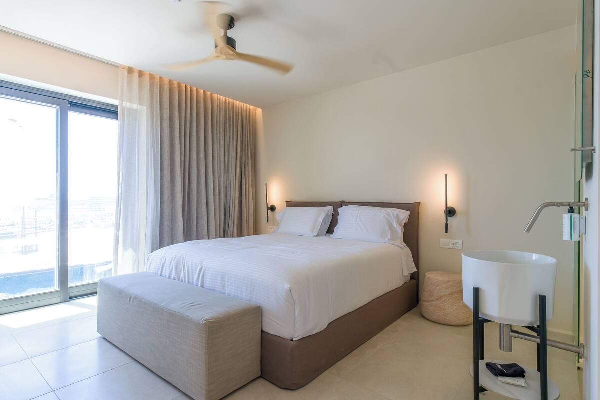 A modern hotel room with a large bed, a bedside table, a small stool, and a bathroom with a sink and mirror.