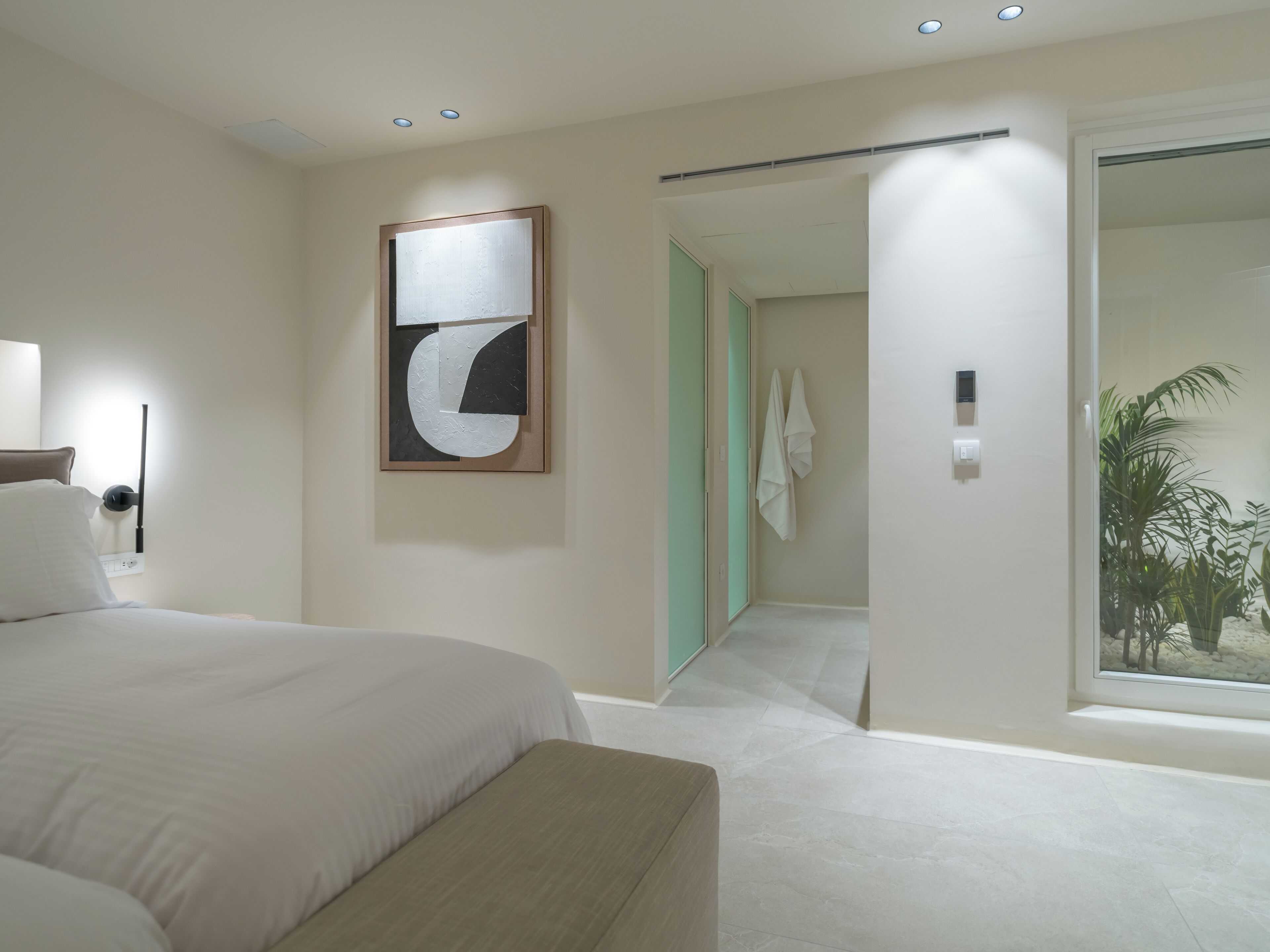 Een moderne hotelkamer met een groot bed, een wanddecoratie met abstracte kunst, een badkamer met een grote spiegel en uitzicht op een palmboom door het raam.