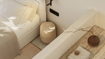 A modern, minimalist bedroom with a bed, a round wooden stool, a wall-mounted light, and a small wooden table with a towel and a hairbrush.