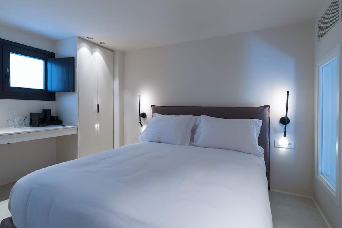 A neatly made bed in a hotel room with a bedside table, a desk, and a window.