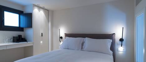 A neatly made bed in a hotel room with a bedside table, a desk, and a window.