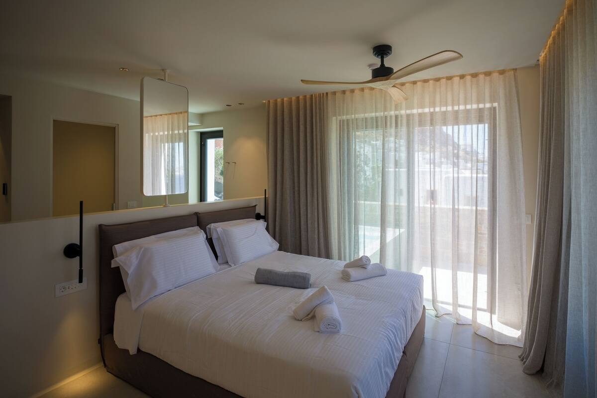 A modern hotel bedroom with a large bed, a ceiling fan, and sheer curtains allowing natural light.