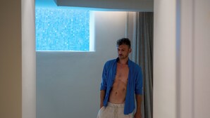 Un hombre con camisa azul y pantalones cortos beige parado en un pasillo con ventana y cortina.