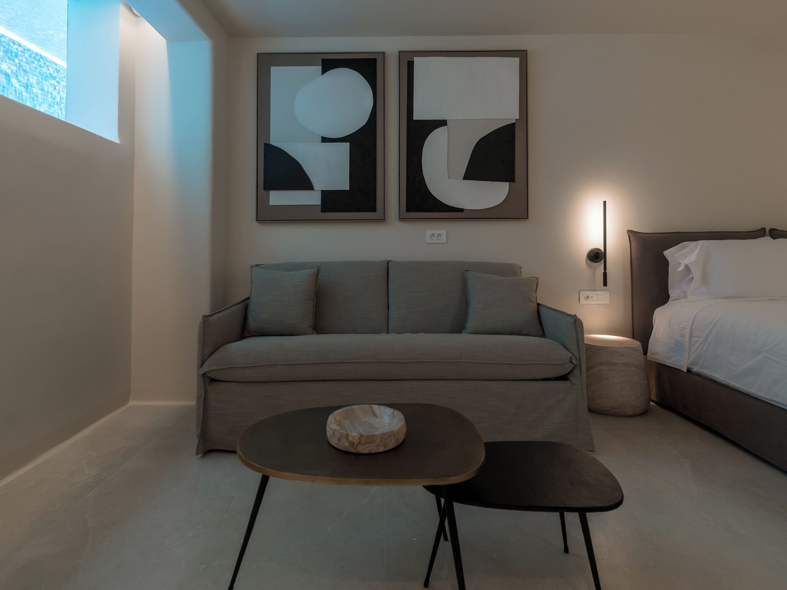A modern bedroom with a grey sofa, a small round table, and a bed with white linens. There are abstract paintings on the wall and a window providing natural light.