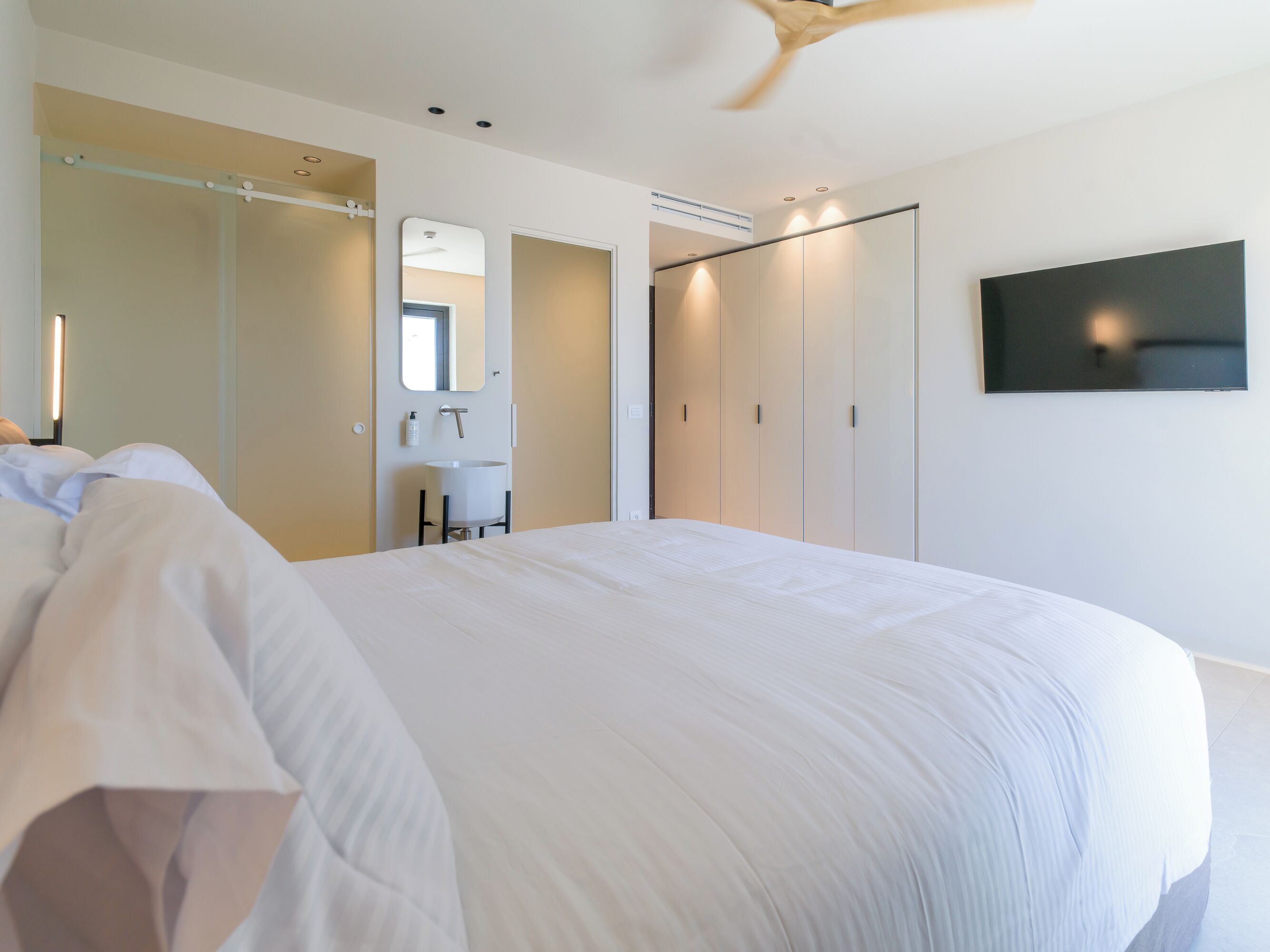 Une chambre moderne avec un grand lit, une armoire intégrée, un miroir, un téléviseur fixé au mur et un ventilateur de plafond.