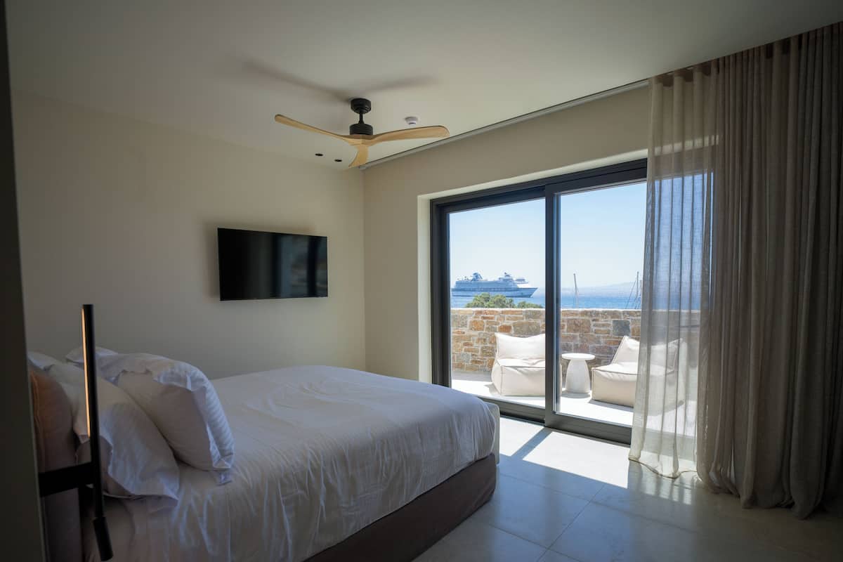 A bedroom with a large bed, a ceiling fan, a TV mounted on the wall, and a view of the sea through a sliding glass door.