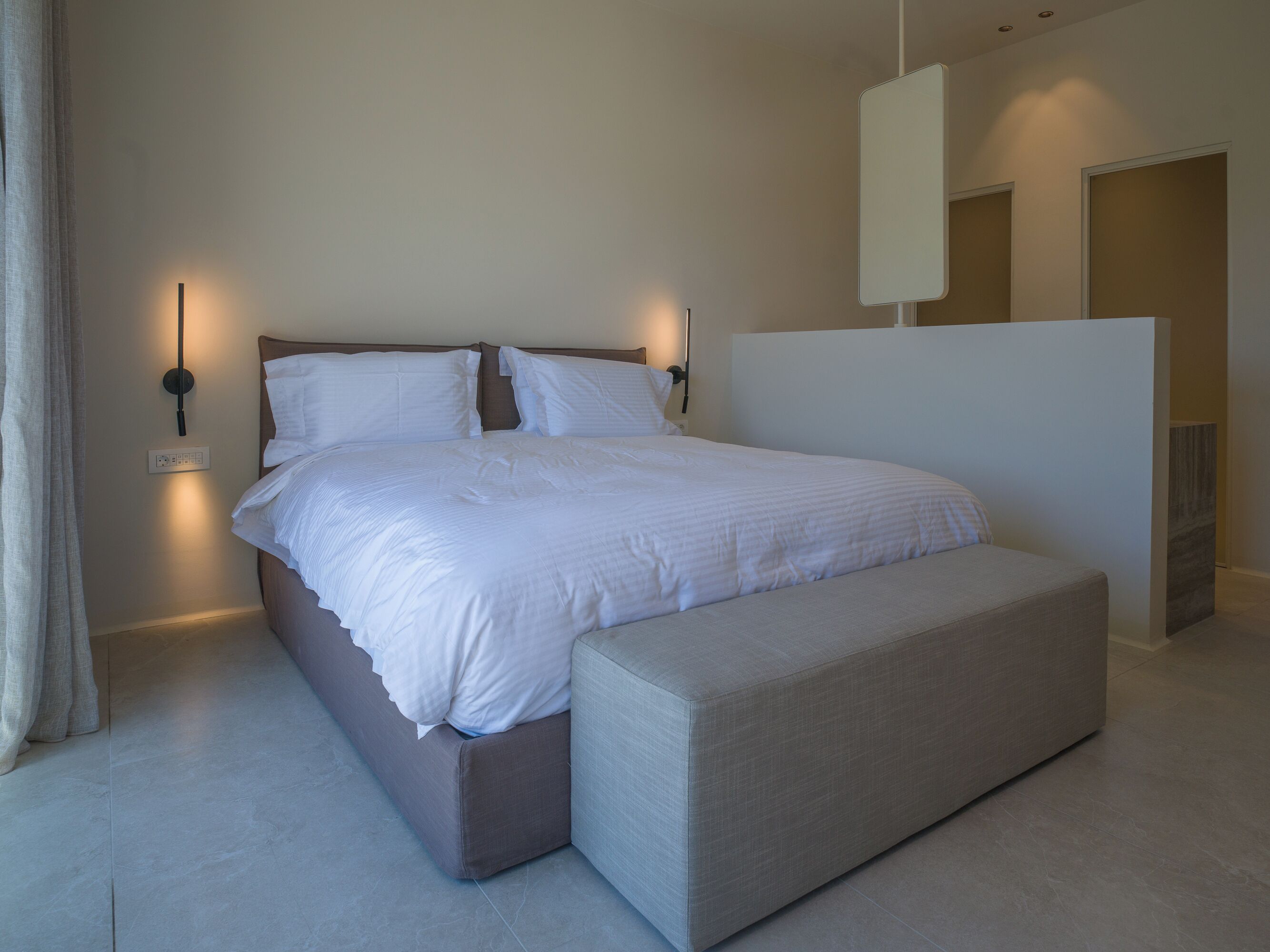 A modern bedroom with a large bed, bedside tables, a bench, and a wall-mounted light fixture.