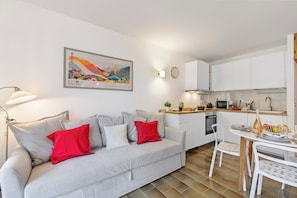A modern living room with a grey sofa, red pillows, a white coffee table, and a kitchen area with white cabinets and a microwave.