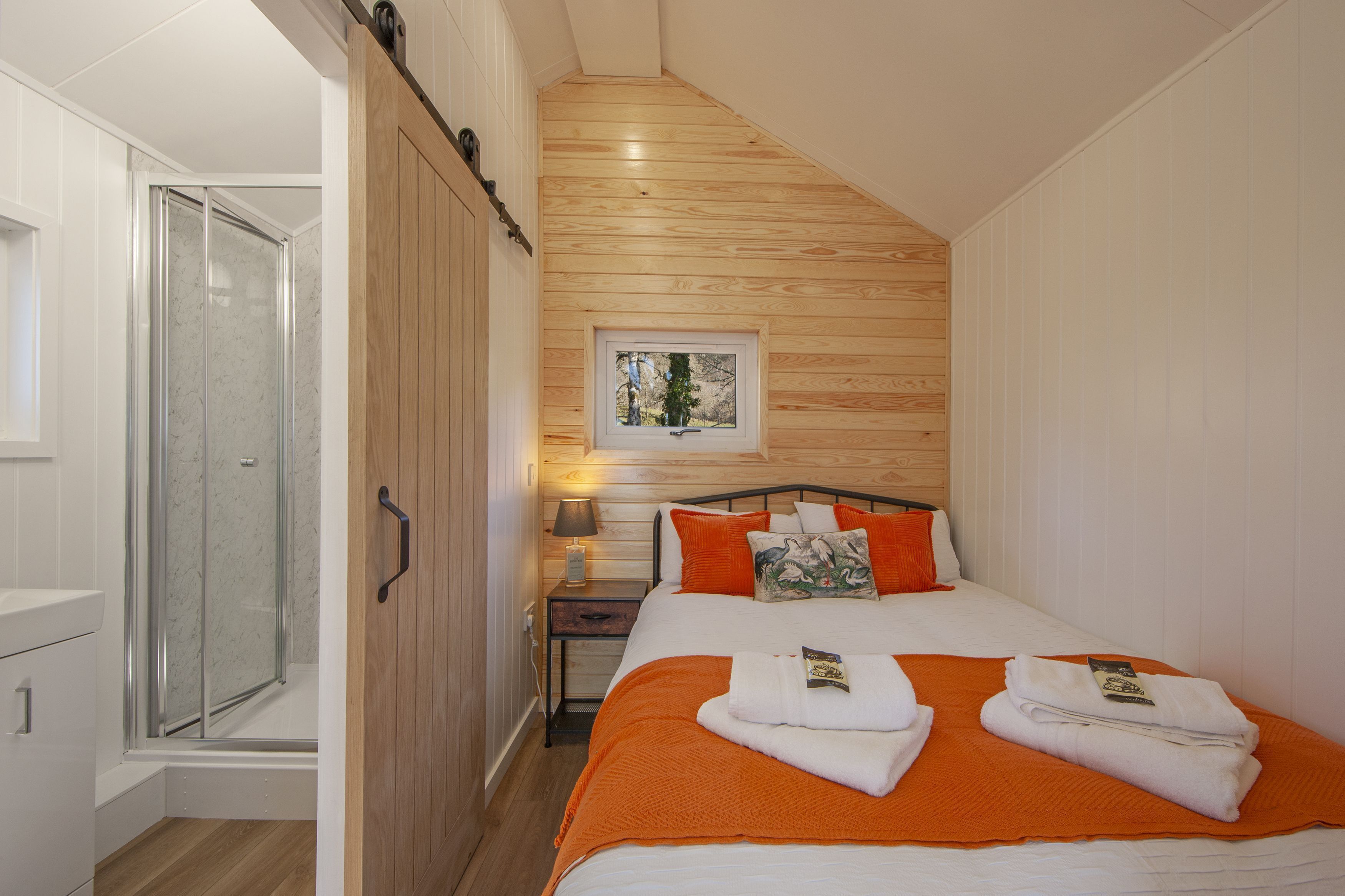 Un dormitorio con techo de madera, una cama con ropa de cama naranja, una mesita de noche con lámpara y una ventana con vistas a árboles.