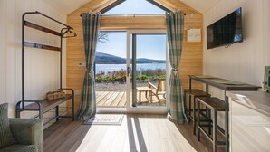 Une pièce au décor intérieur en bois, avec une grande fenêtre ornée de rideaux, un téléviseur fixé au mur et une vue sur un lac et des montagnes.