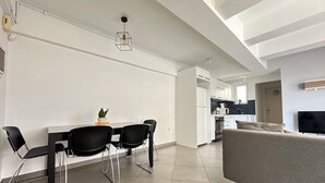 Een moderne woonkamer met een grijze bank, een eettafel met zwarte stoelen, en een keuken op de achtergrond.