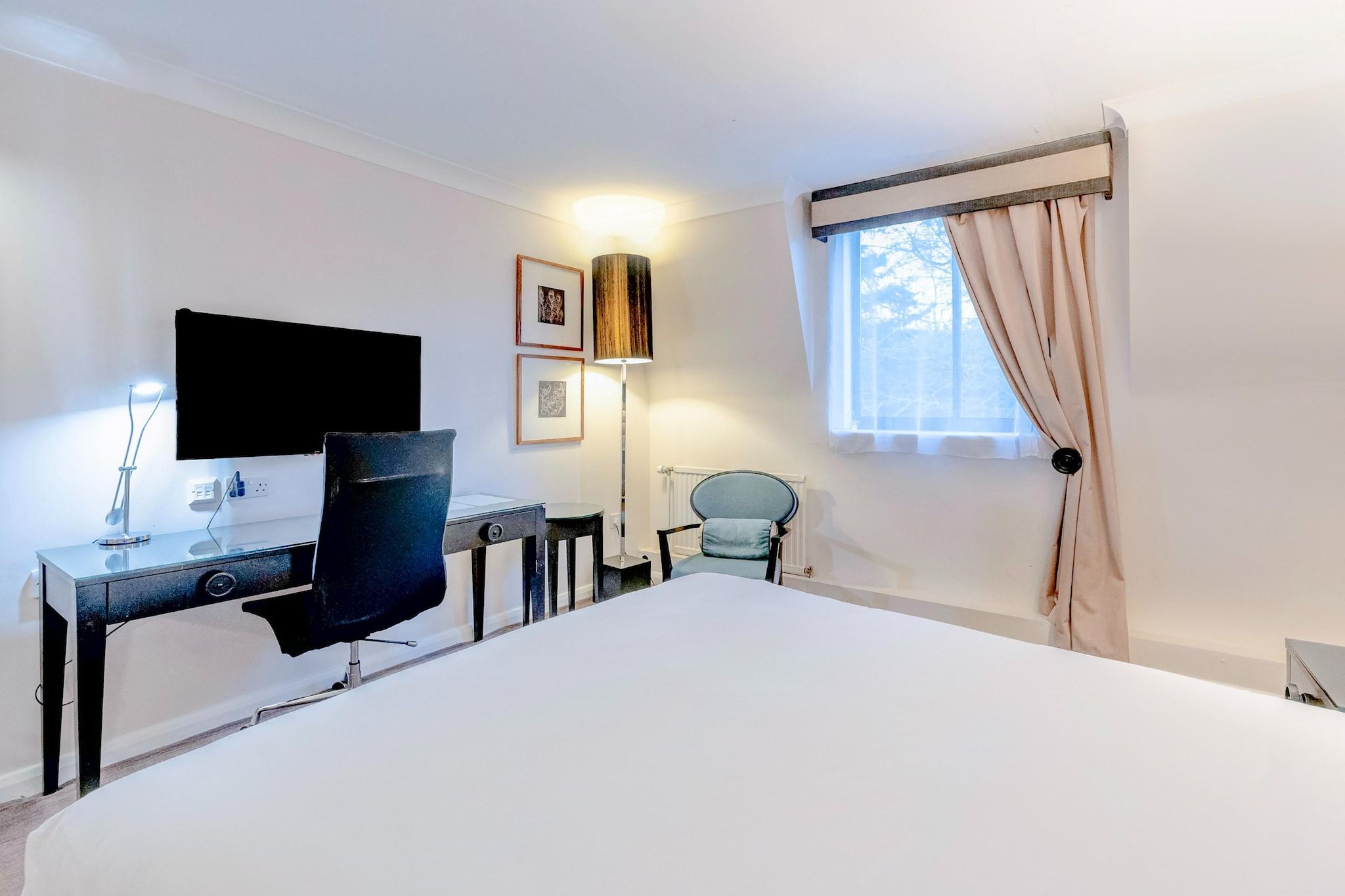 A hotel room with a large bed, a desk with a chair, a television, a lamp, and a window with curtains.