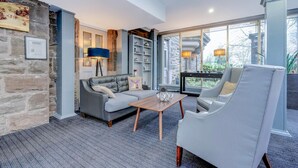 A modern living room with a stone wall, a grey sofa, a white armchair, a wooden coffee table, and a bookshelf. - The Landmark Hotel and Leisure Club | SUPR DBL (Dundee)