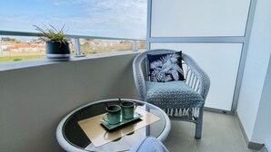 Un balcone con vista sul cielo e edifici in lontananza, un tavolino rotondo con un vassoio e due tazze, una sedia con cuscino decorato e una pianta in vaso.