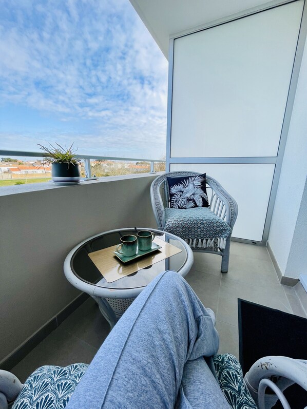 Un balcón con vista al cielo y edificios a lo lejos, una mesa redonda con una bandeja y dos tazas, una silla con cojín estampado y una planta en maceta.