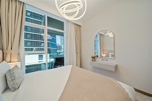 A modern hotel room with a large window, a bed with a textured blanket, a bedside table with a lamp, and a mirror with a decorative frame. - Modern 2BR – Canal-Side Elegance (Dubai)