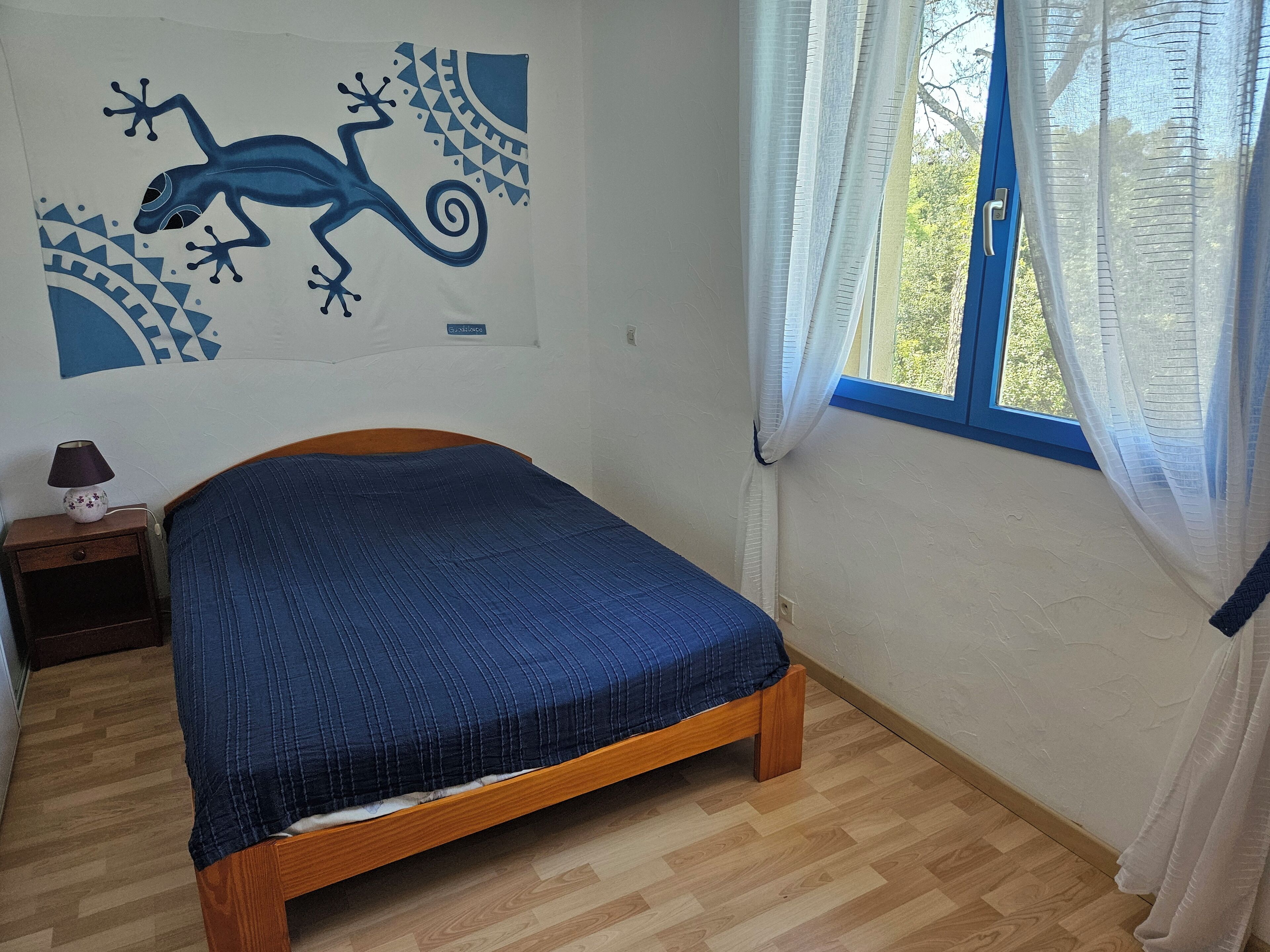 A bedroom with a wooden bed, a nightstand with a lamp, a window with sheer curtains, and a wall mural.
