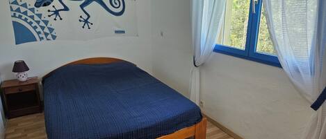 A bedroom with a wooden bed, a nightstand with a lamp, a window with sheer curtains, and a wall mural.