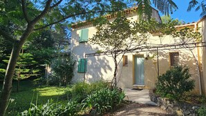 Una casa de dos pisos con fachada de piedra, contraventanas verdes y un jardín con diversas plantas y un sendero que conduce a la entrada.