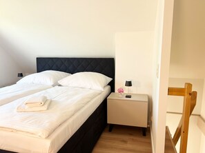 Ein ordentlich bezogenes Bett mit weißen Leinen und einem gefalteten Handtuch, ein schwarzer möbelbezogener Kopfteil, ein Nachttisch mit Lampe und einem Blumenstrauß sowie ein an der Wand angelehnt stehender Holzhocker.